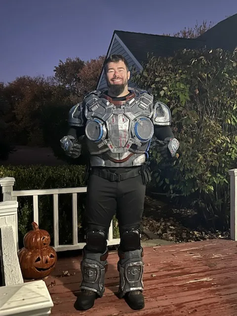 A man in a full-body, heavily armored sci-fi costume stands on a wooden porch at dusk, smiling. The costume includes a chest…