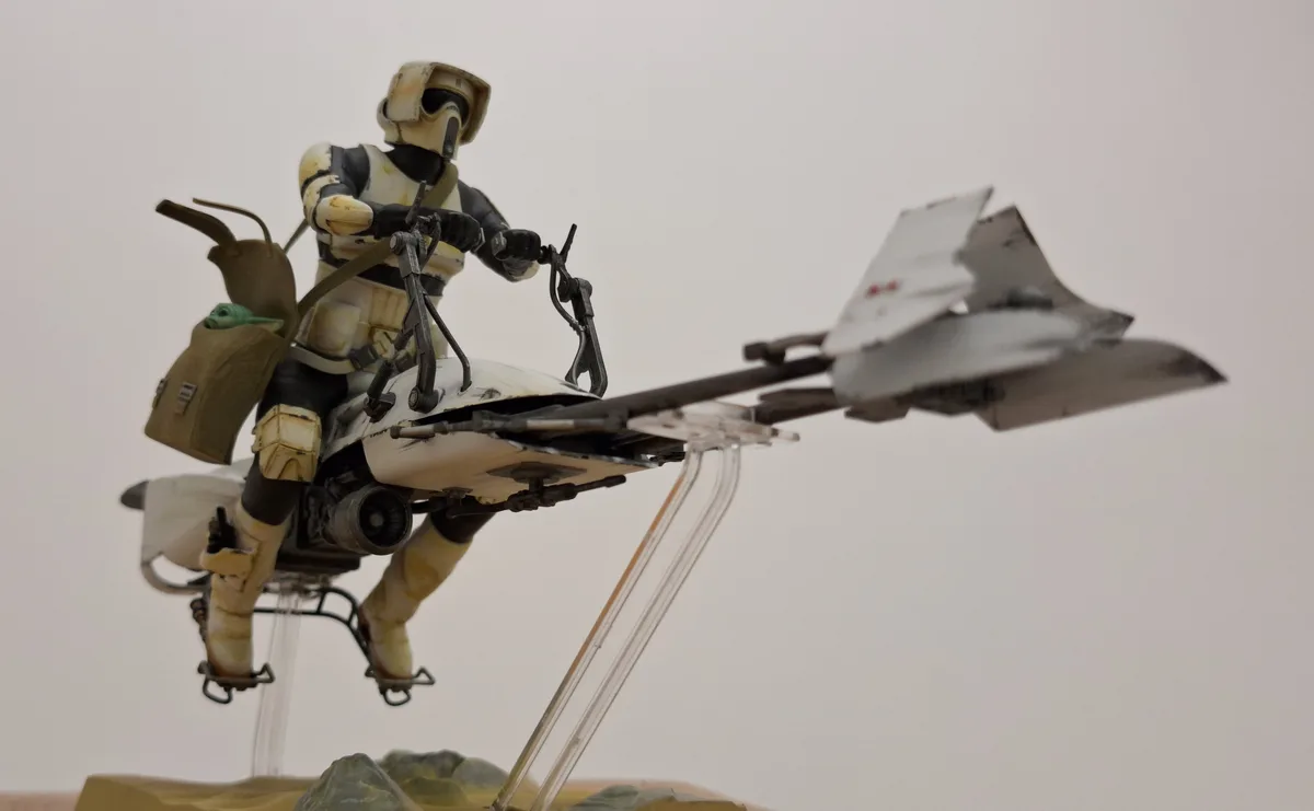 A Star Wars action figure of a Mandalorian rider in weathered white armor, mounted on a white podracer with a small green…