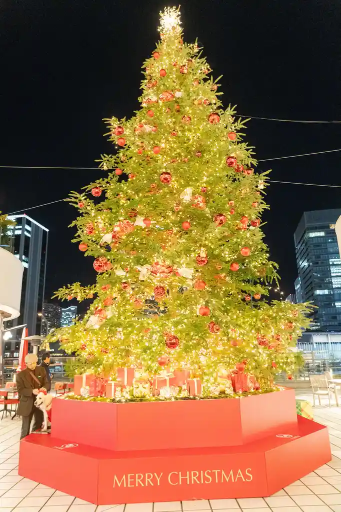 A large, brightly lit Christmas tree adorned with red ornaments and white lights stands on a red pedestal with 'MERRY…