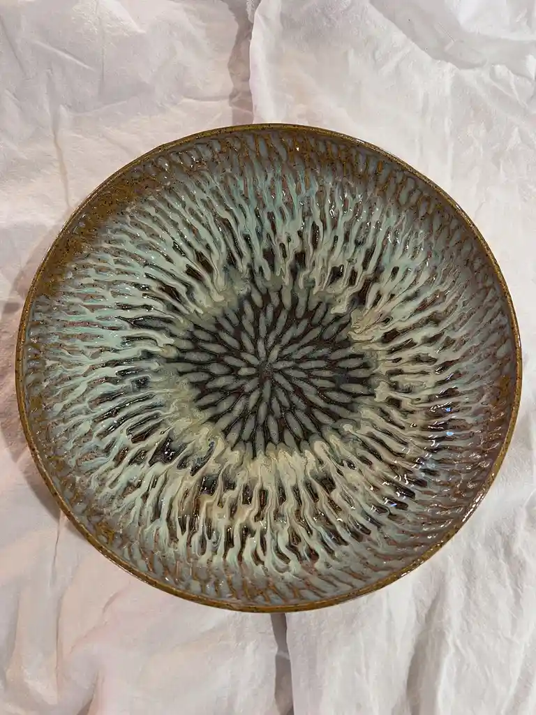 A top-down photo of a ceramic bowl with a radial, sunburst-like glaze pattern in brown and teal, resting on a white cloth…