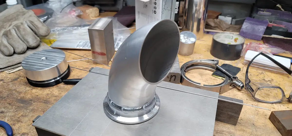 A workshop scene shows a shiny metal 90-degree elbow pipe resting on a metal block, surrounded by tools like gloves, tape…