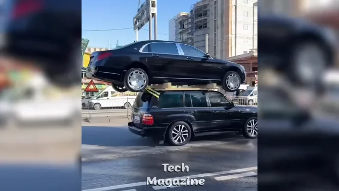 A black sedan is visibly balanced on top of a black station wagon driving on a city street, with buildings and other…