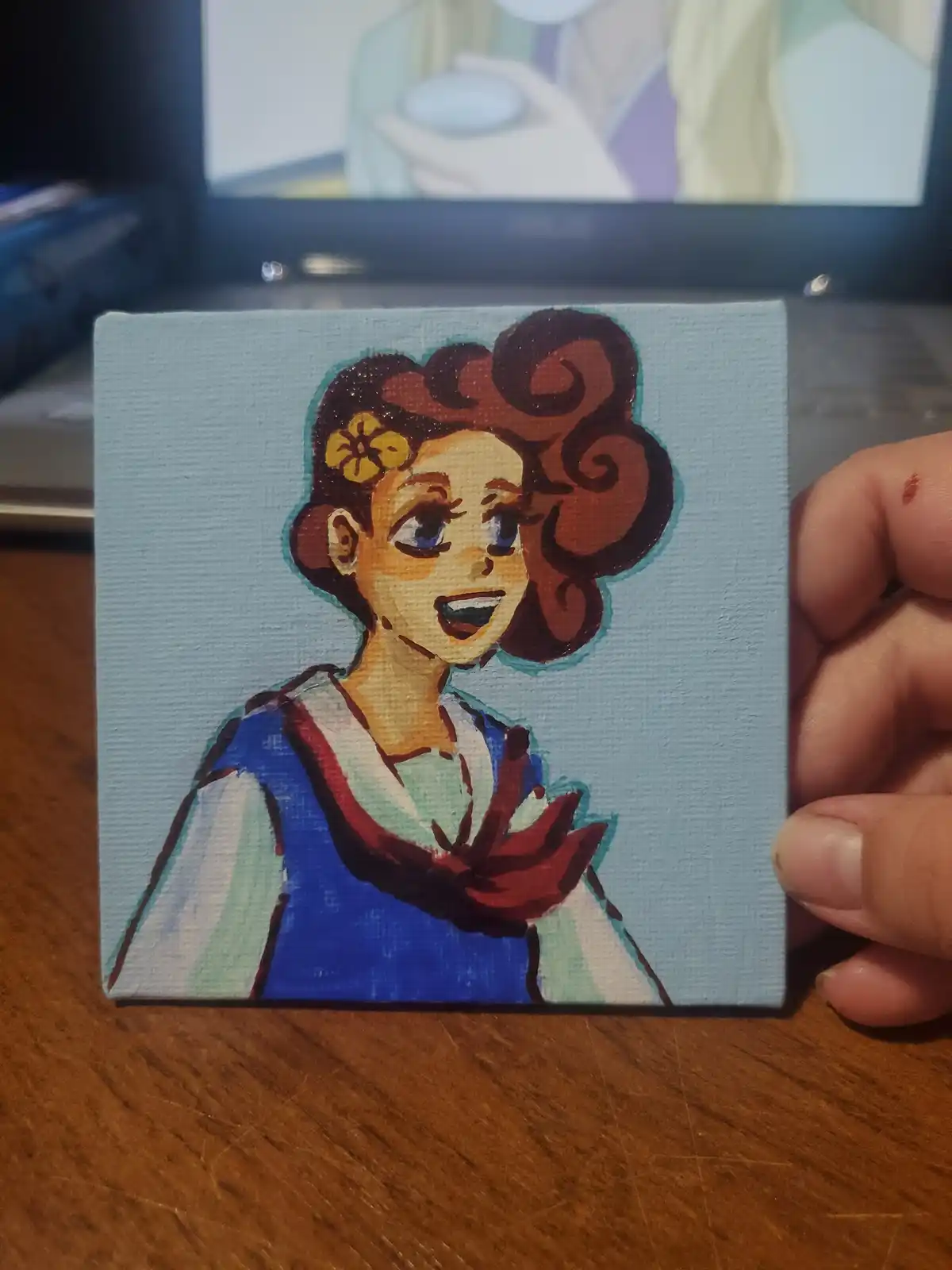 A hand holds a small painted canvas depicting a cheerful anime-style girl with brown curly hair, a yellow flower in her…