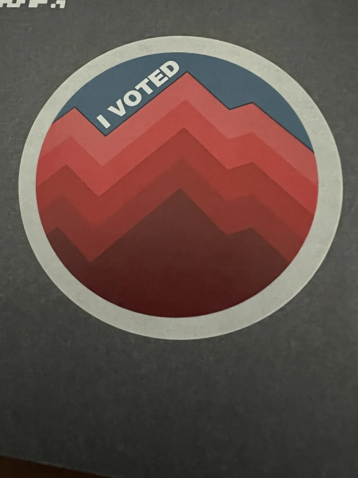 A circular 'I VOTED' sticker with a stylized mountain range design in red and blue, placed on a dark gray surface. The text…