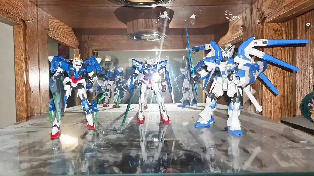 Three blue and white Gundam model kits are displayed on a glass shelf inside a wooden cabinet, with reflections visible on…
