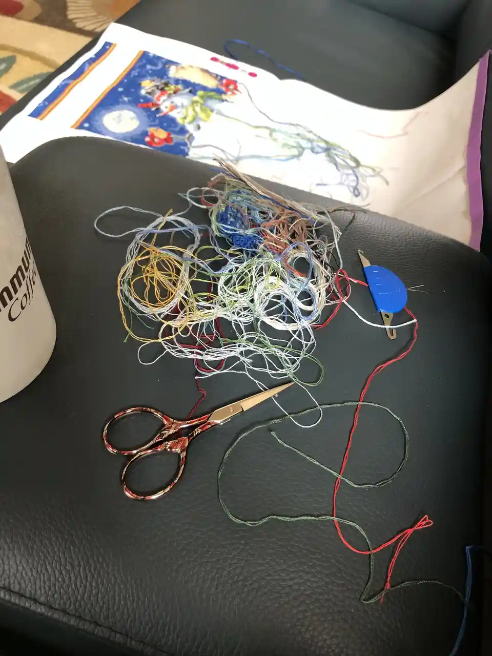 A messy pile of colorful embroidery floss sits on a black leather couch next to a pair of decorative scissors, a blue thread…