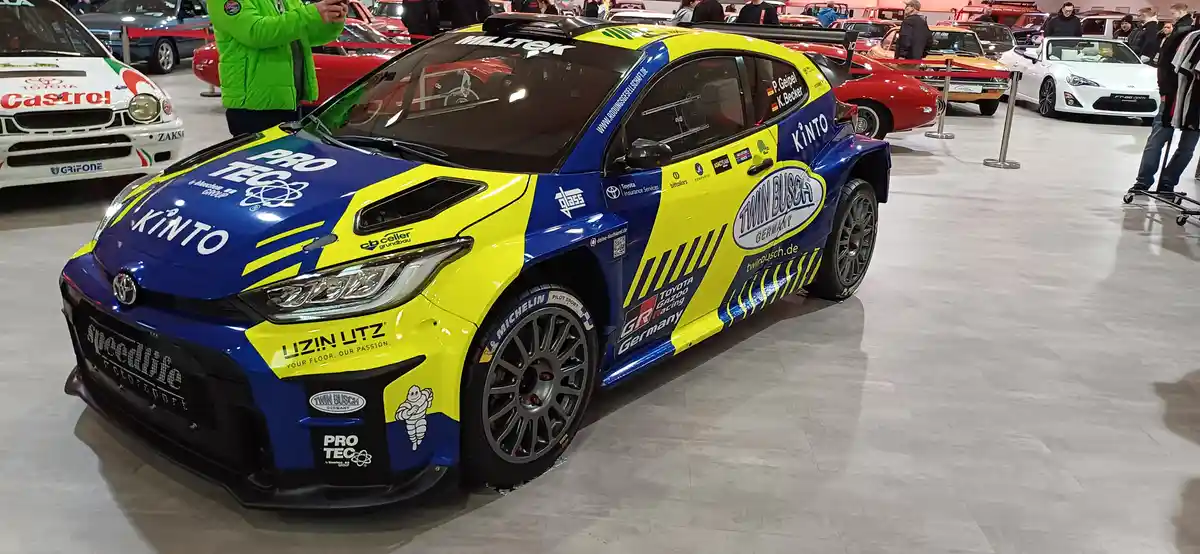 A blue and yellow Toyota GR Yaris Rally1 race car with 'Twin Busch Germany' and 'Kinto' branding is displayed indoors at a…