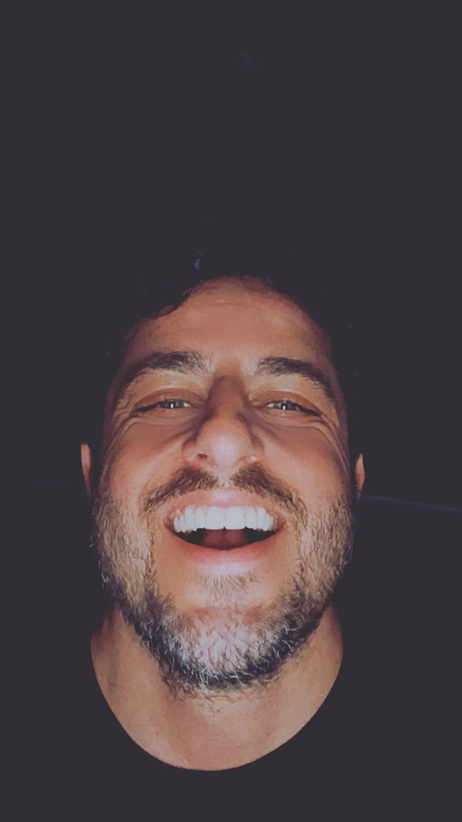 A close-up photo of a man with dark hair and a beard, laughing heartily with his mouth wide open, showing his teeth, against…