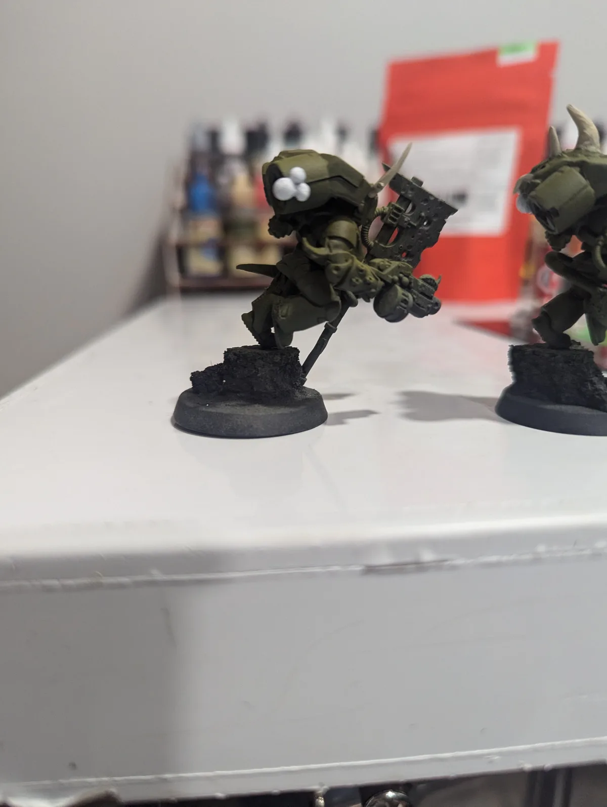 Two green-painted miniature figurines, likely from a tabletop wargame, stand on black bases on a white surface. The figurine…