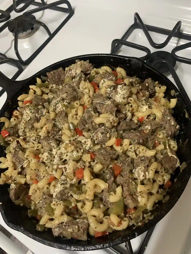 A close-up photo shows a black cast-iron skillet on a white stovetop, filled with elbow macaroni, chunks of browned meat…