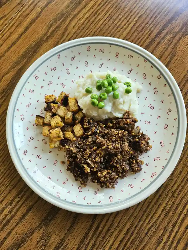 A plate of food on a wooden table contains mashed potatoes topped with green peas, a pile of textured brown vegan meat…