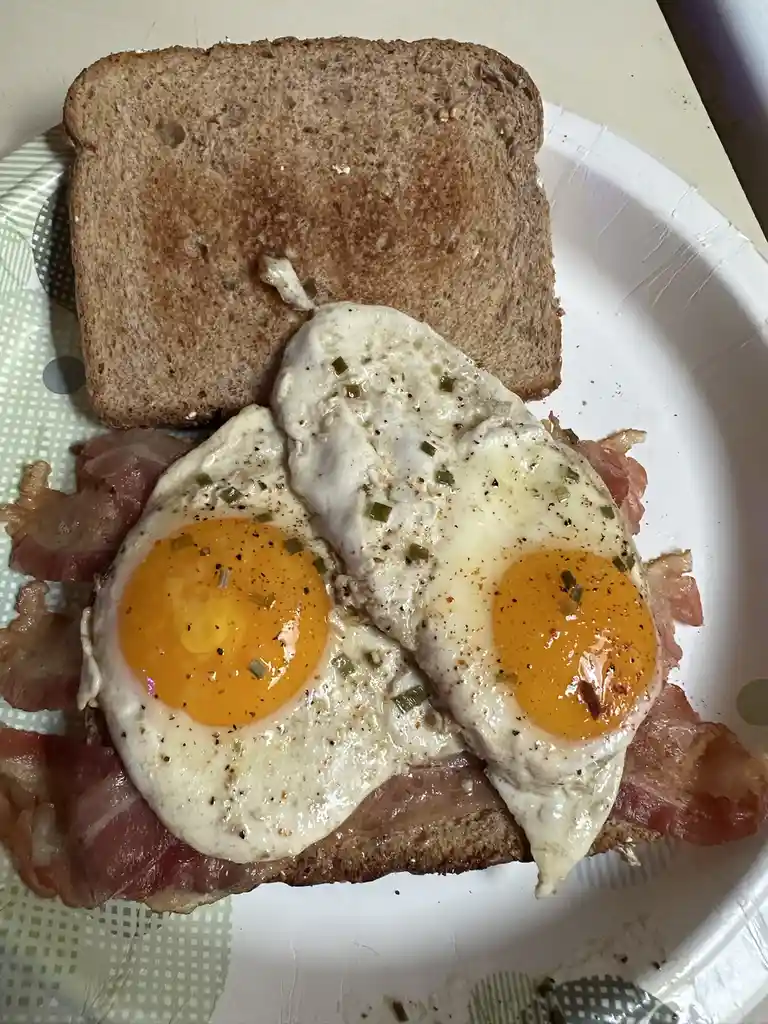 A close-up photo shows a breakfast sandwich on a paper plate, featuring two sunny-side-up eggs with bright yellow yolks…