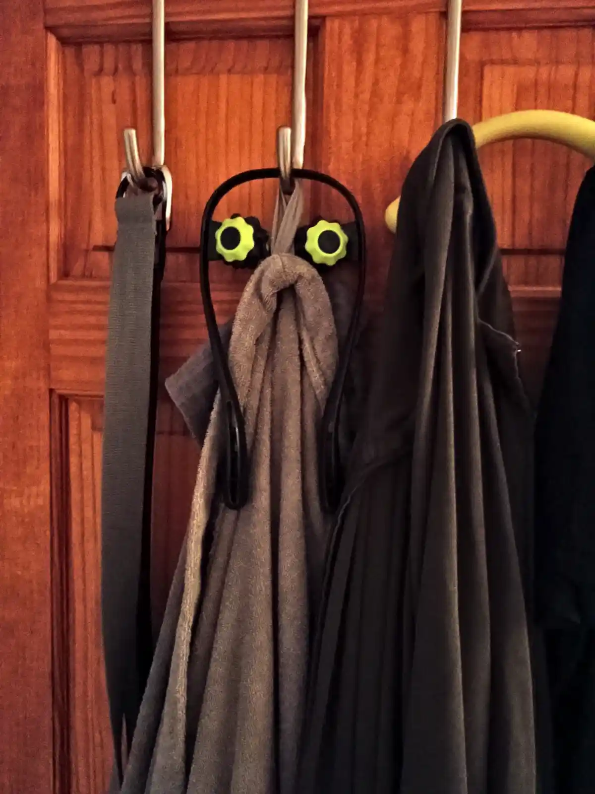A brown towel hangs on a hook, fashioned into a ghost costume with two bright green, flower-shaped knobs serving as eyes…