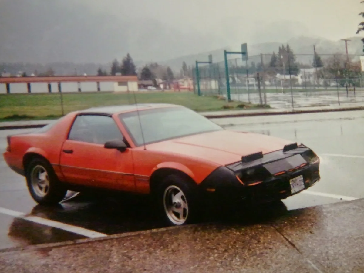 A vintage orange and black two-door sports car, likely a Ford Mustang, is parked in a wet parking lot on an overcast day…