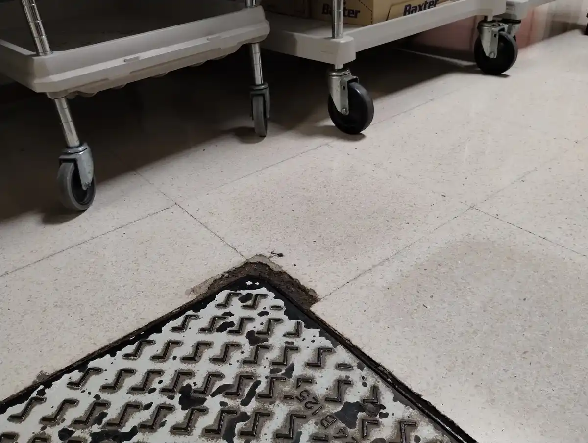 A close-up photo shows a dirty, triangular manhole cover partially embedded in a light-colored tiled floor, with the edges…