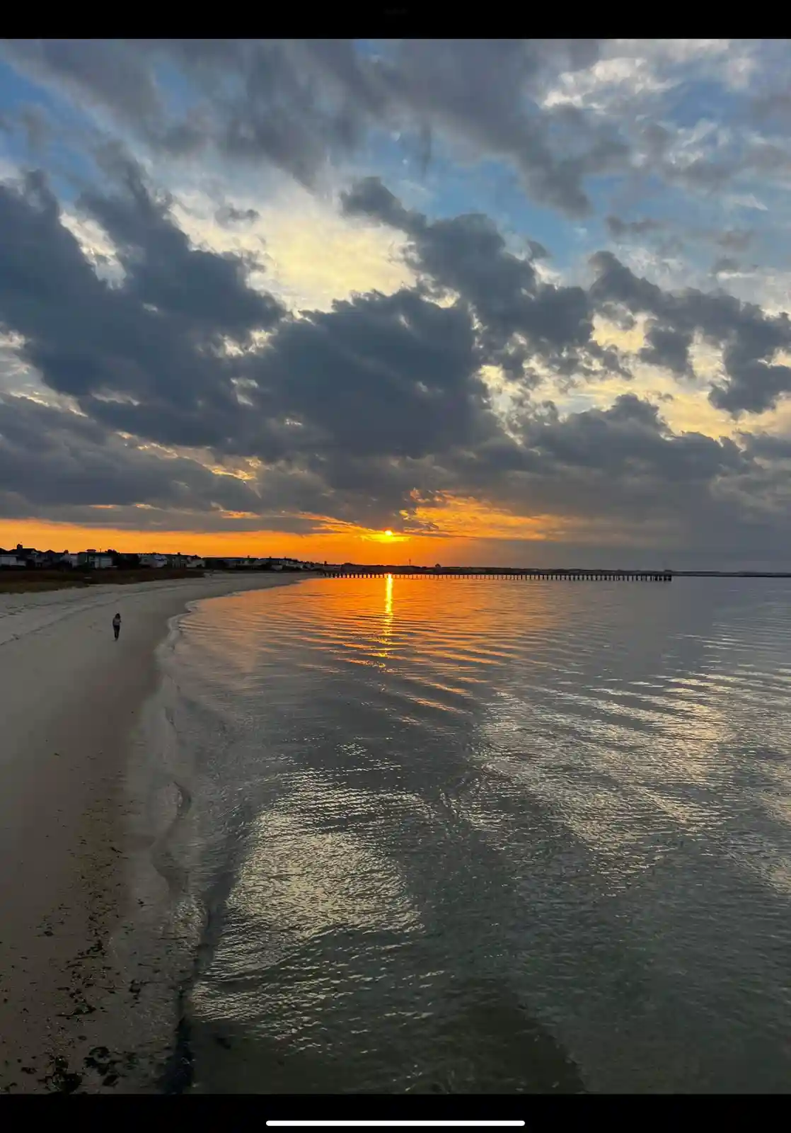 A serene beach sunset photo with a lone person walking along the shoreline, the sun setting on the horizon casting orange…