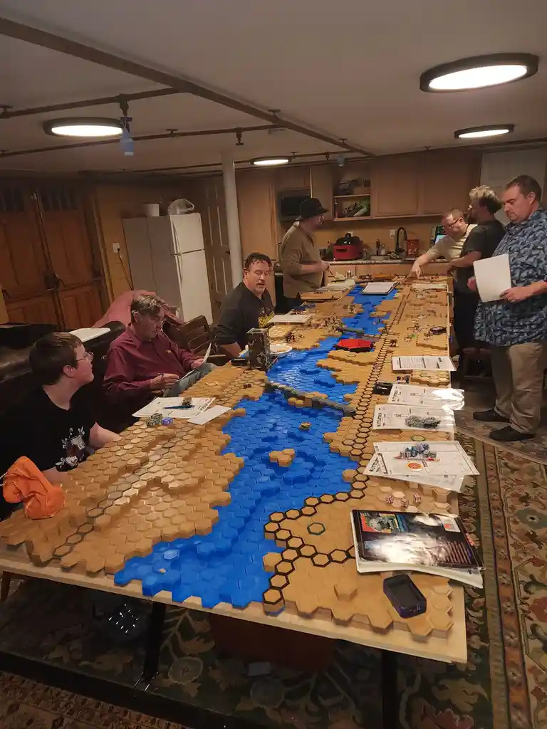 A group of men are gathered around a very long table in a basement, playing a large-scale tabletop wargame using hexagonal…
