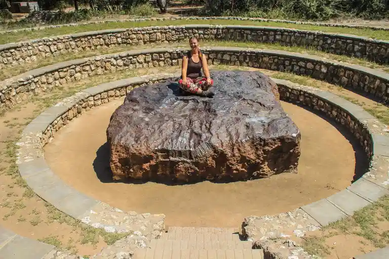 A woman sits cross-legged on a large, dark, textured fossilized rock in the center of a circular stone enclosure. The rock…