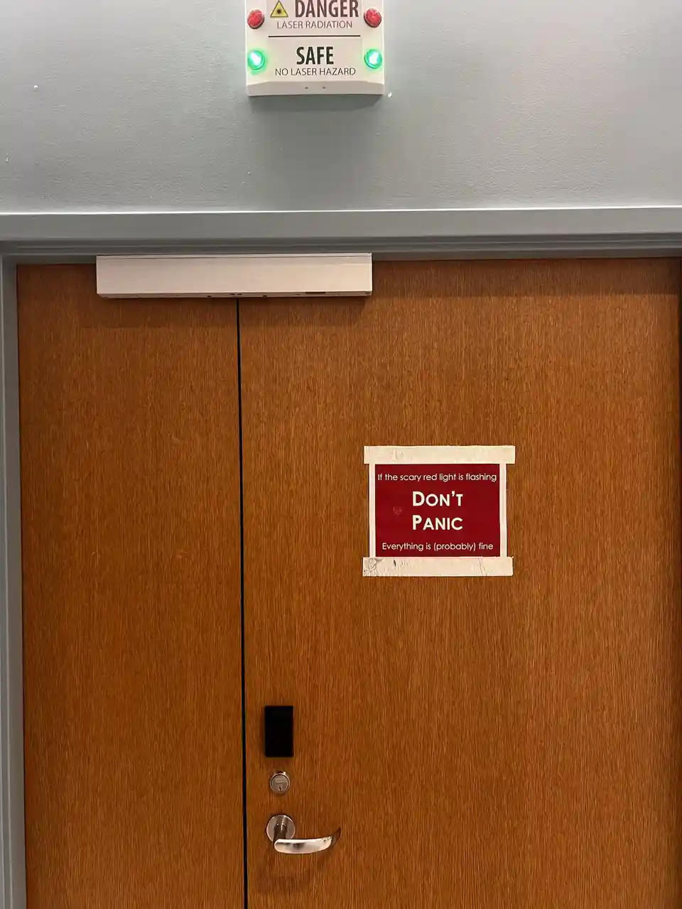 A wooden door has a red sign taped to it that reads 'If the scary red light is flashing DON'T PANIC Everything is (probably)…