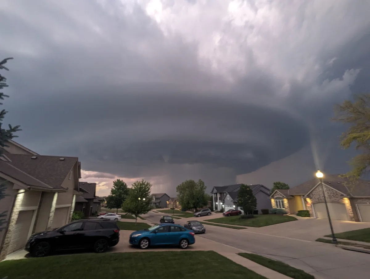 A suburban neighborhood street at dusk under a massive, ominous, dark storm cloud that appears to be looming directly over…