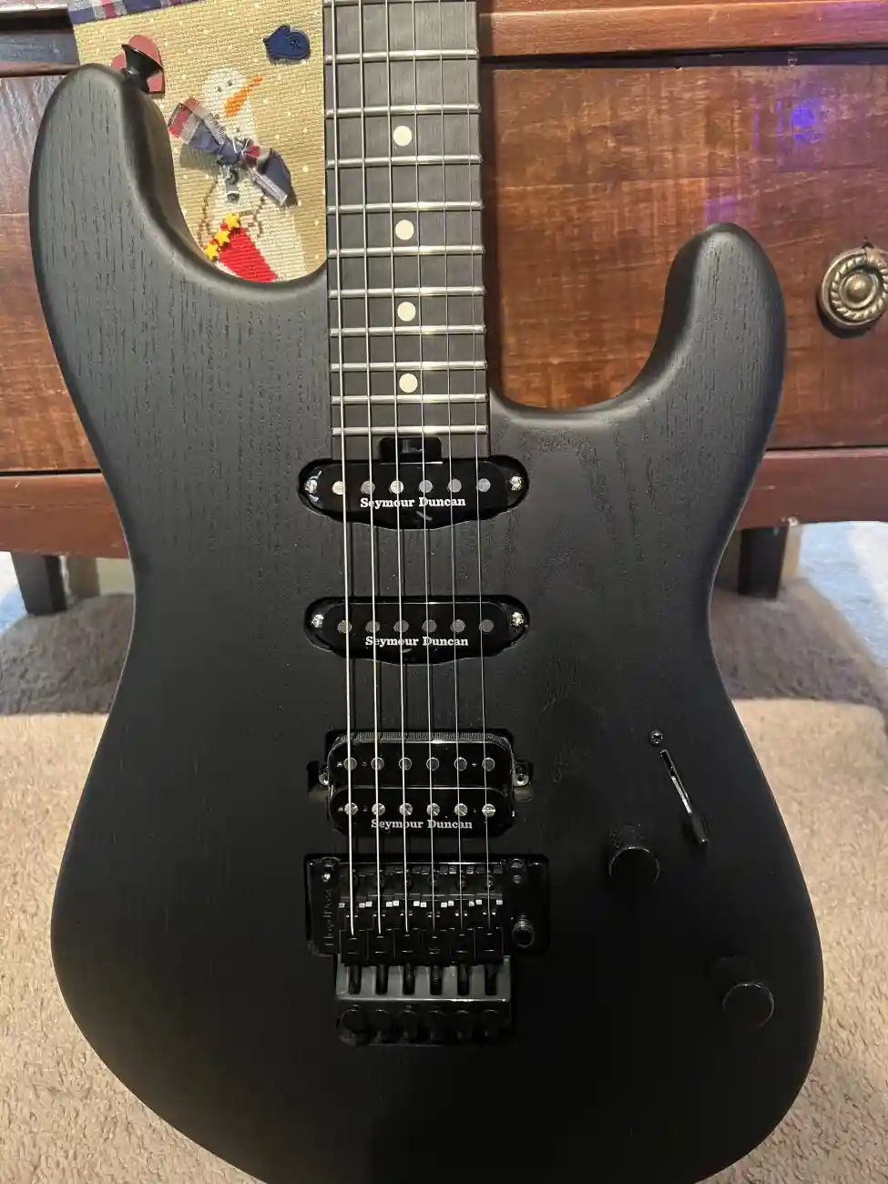 A close-up photo of a black electric guitar with three Seymour Duncan pickups, resting on a carpeted floor in front of a…