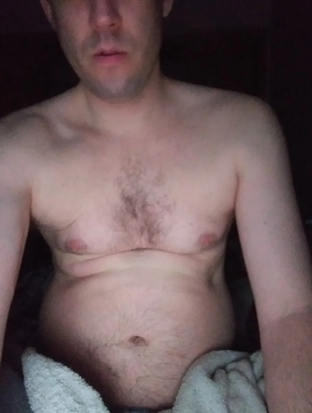 A shirtless man with a hairy chest and belly, sitting with a towel draped over his lower body, looking directly at the…