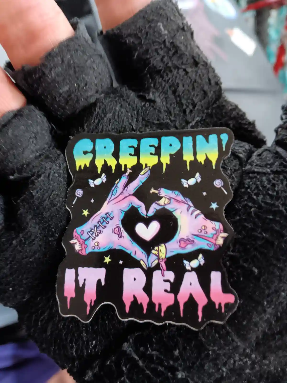 A person holds a black sticker with two cartoonish, zombie-like hands forming a heart shape around a white heart. The top…