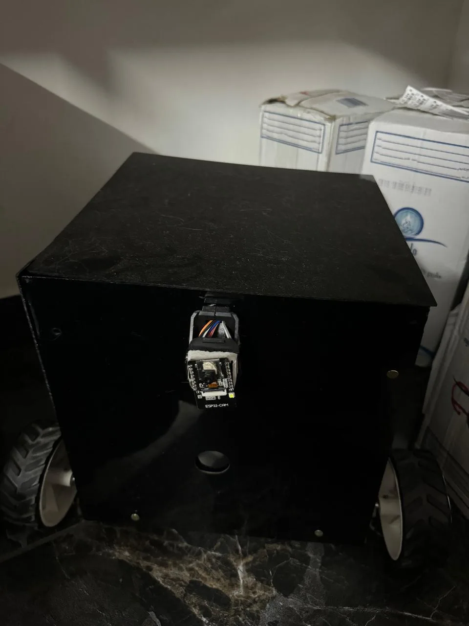 A black box with wheels, resembling a robot, has an esp32-cam module taped to its side with wires exposed. The camera module…