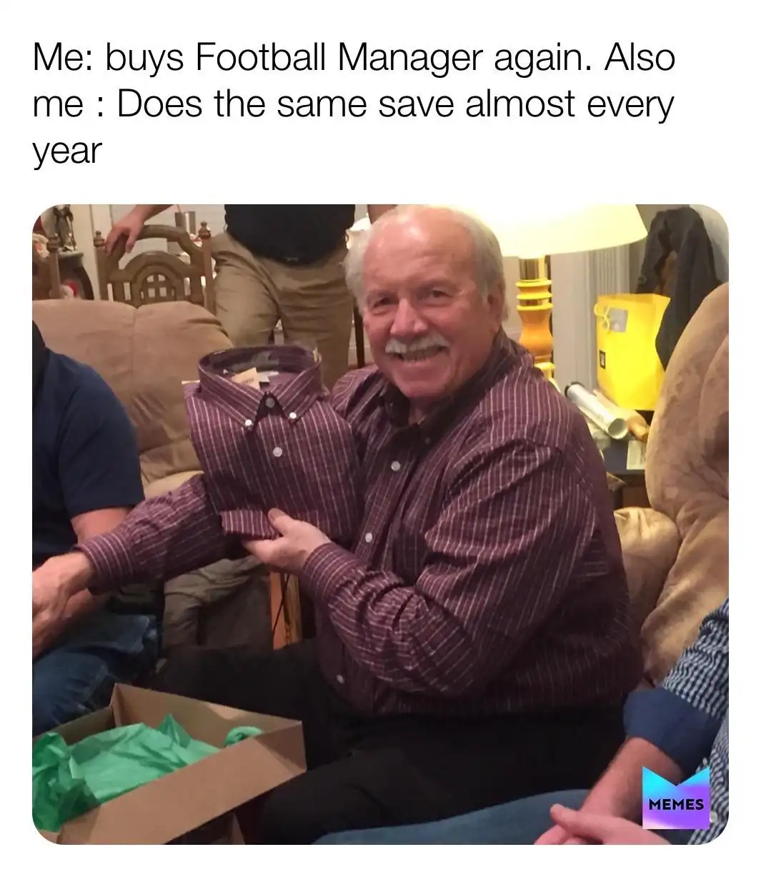 A smiling older man with a mustache, wearing a maroon plaid shirt, holds up a folded identical shirt from a cardboard box…