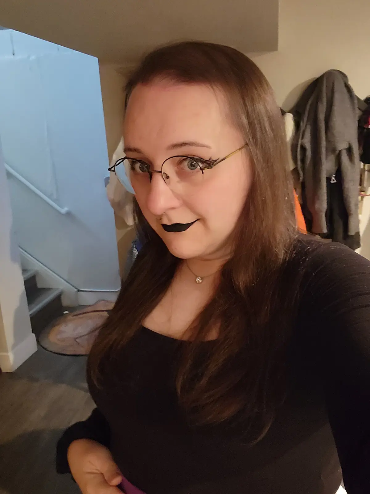 A woman with long brown hair, glasses, and black lipstick poses indoors, looking at the camera with a slight smirk. Behind…