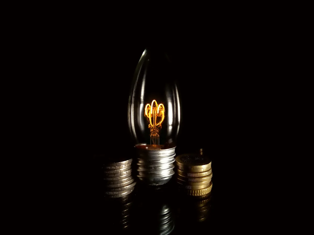 A lit, vintage-style light bulb with a visible filament stands upright on a reflective surface, flanked by two stacks of…