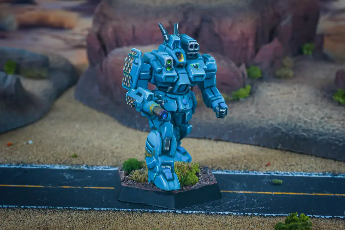 A blue-painted, detailed miniature robot figurine stands on a hexagonal base with small green bushes, positioned on a…