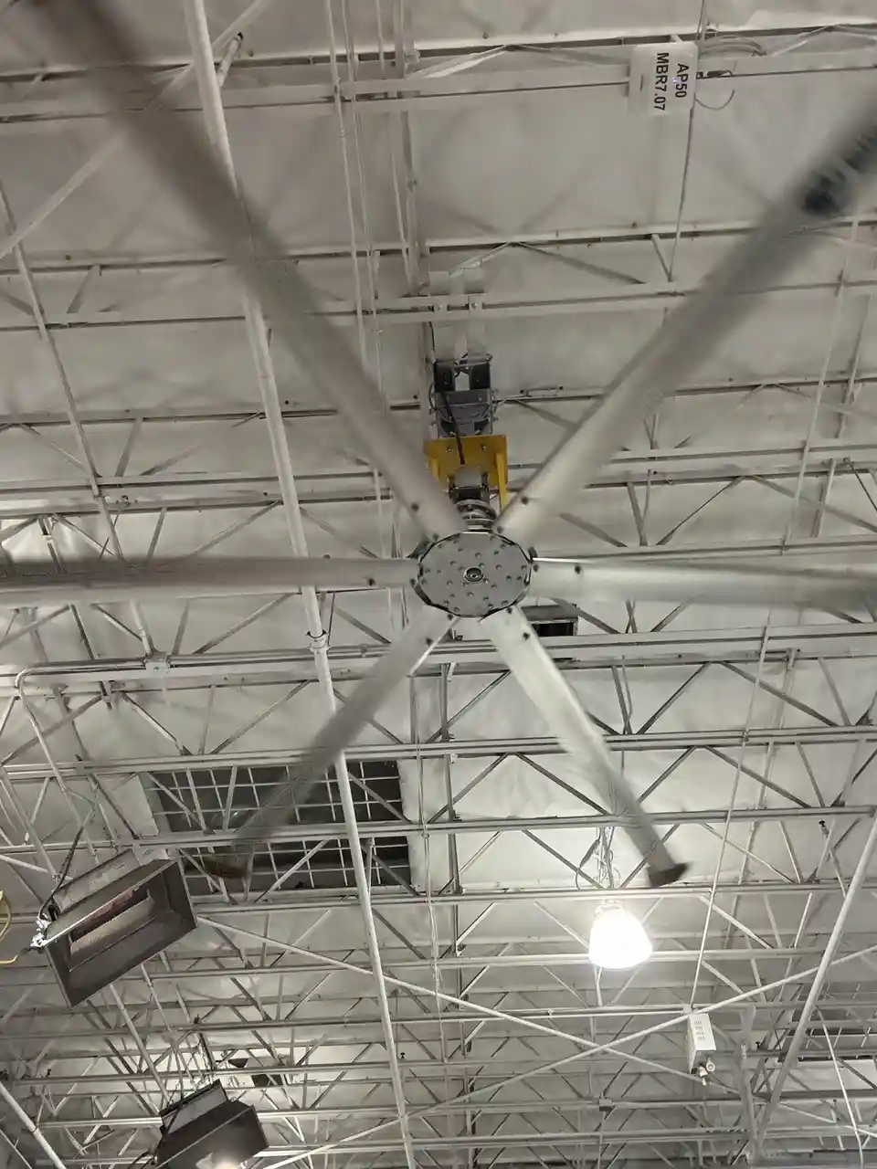 A low-angle photo looking up at a large, industrial-style ceiling fan with blurred blades, mounted on a white metal truss…