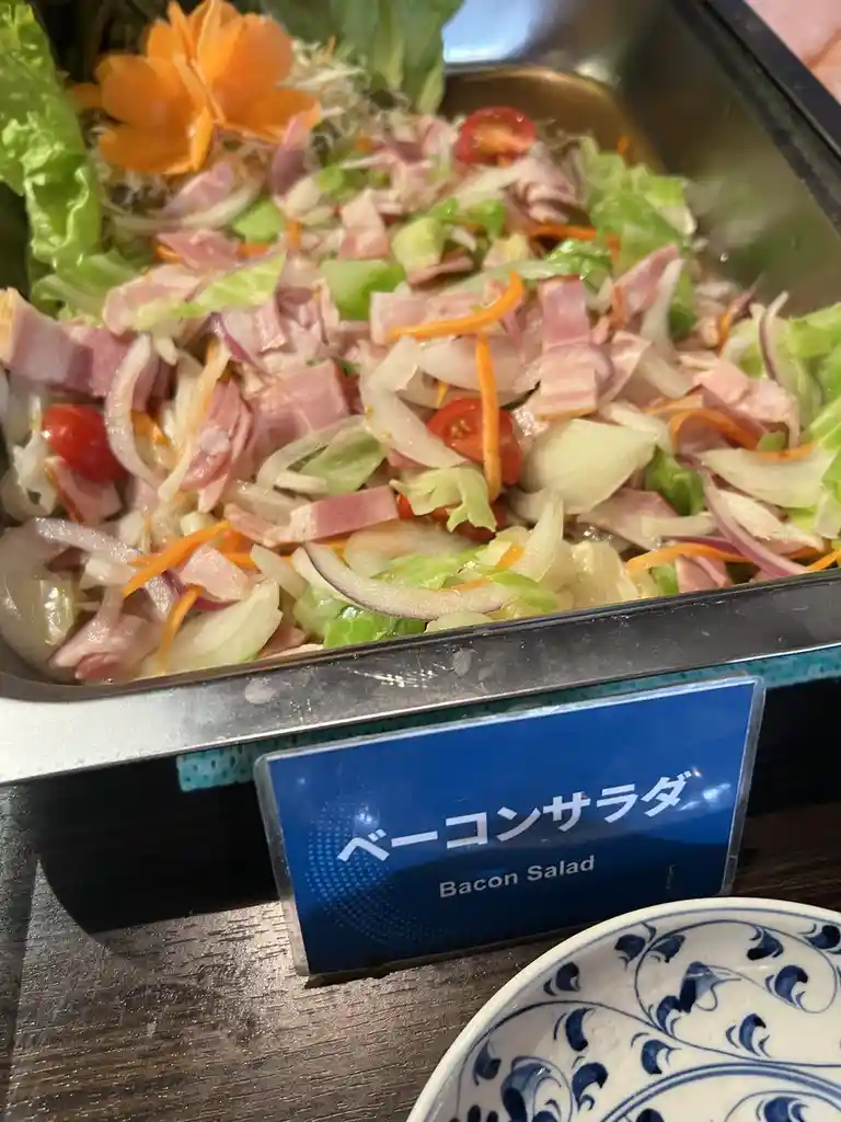 A close-up of a buffet-style salad in a metal tray, featuring lettuce, sliced onions, cherry tomatoes, shredded carrots, and…