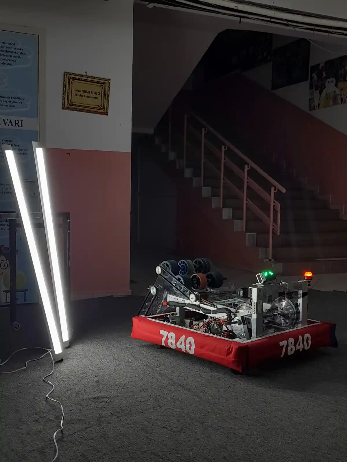 A red robot with exposed mechanical parts and the number 7840 on its sides sits on a carpeted floor next to two tall, bright…