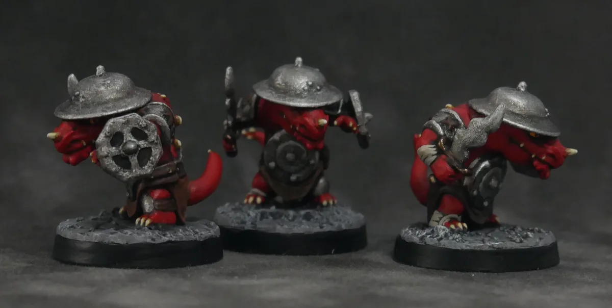 Three red, horned, miniature fantasy creatures wearing silver helmets and armor stand on grey bases against a dark…