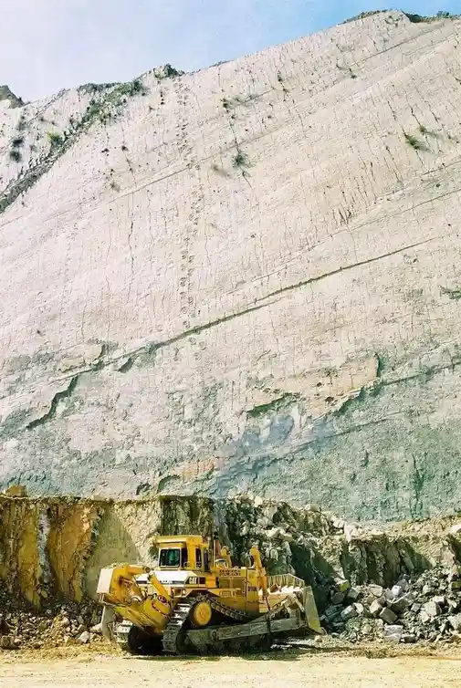 A yellow Caterpillar bulldozer sits at the base of a massive, terraced quarry wall, dwarfed by the sheer scale of the…