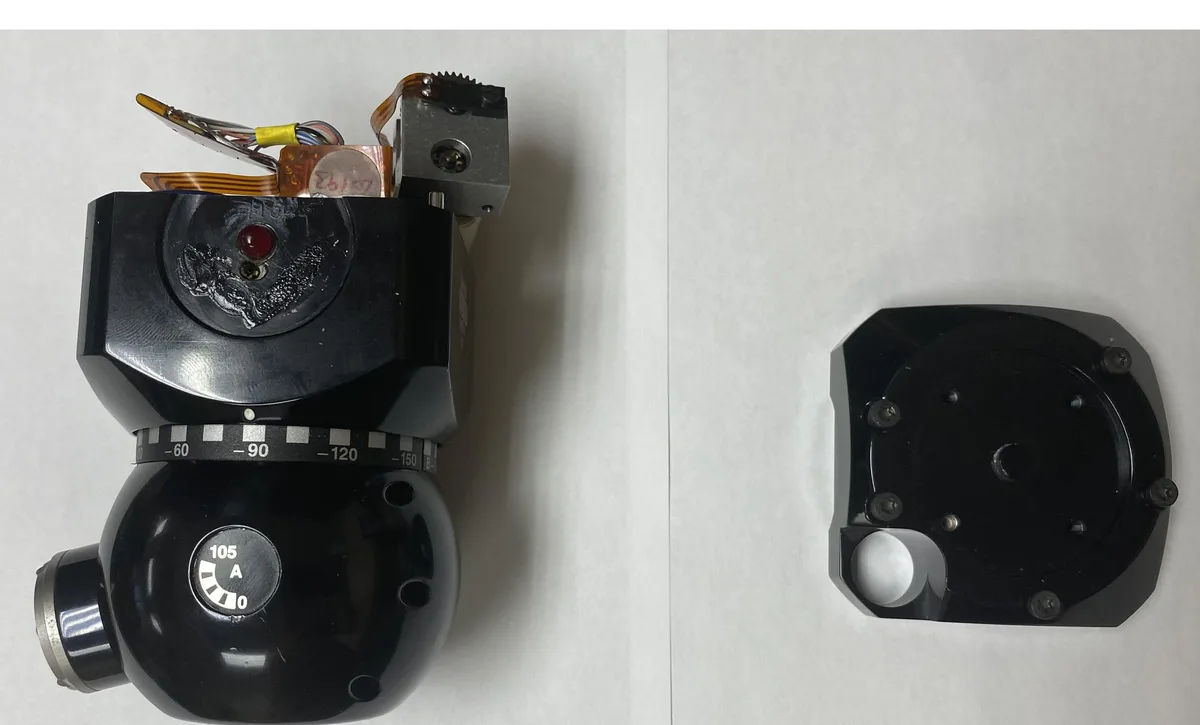 The image shows two black mechanical parts on a white surface. The left part resembles a camera with a red light, a scale…