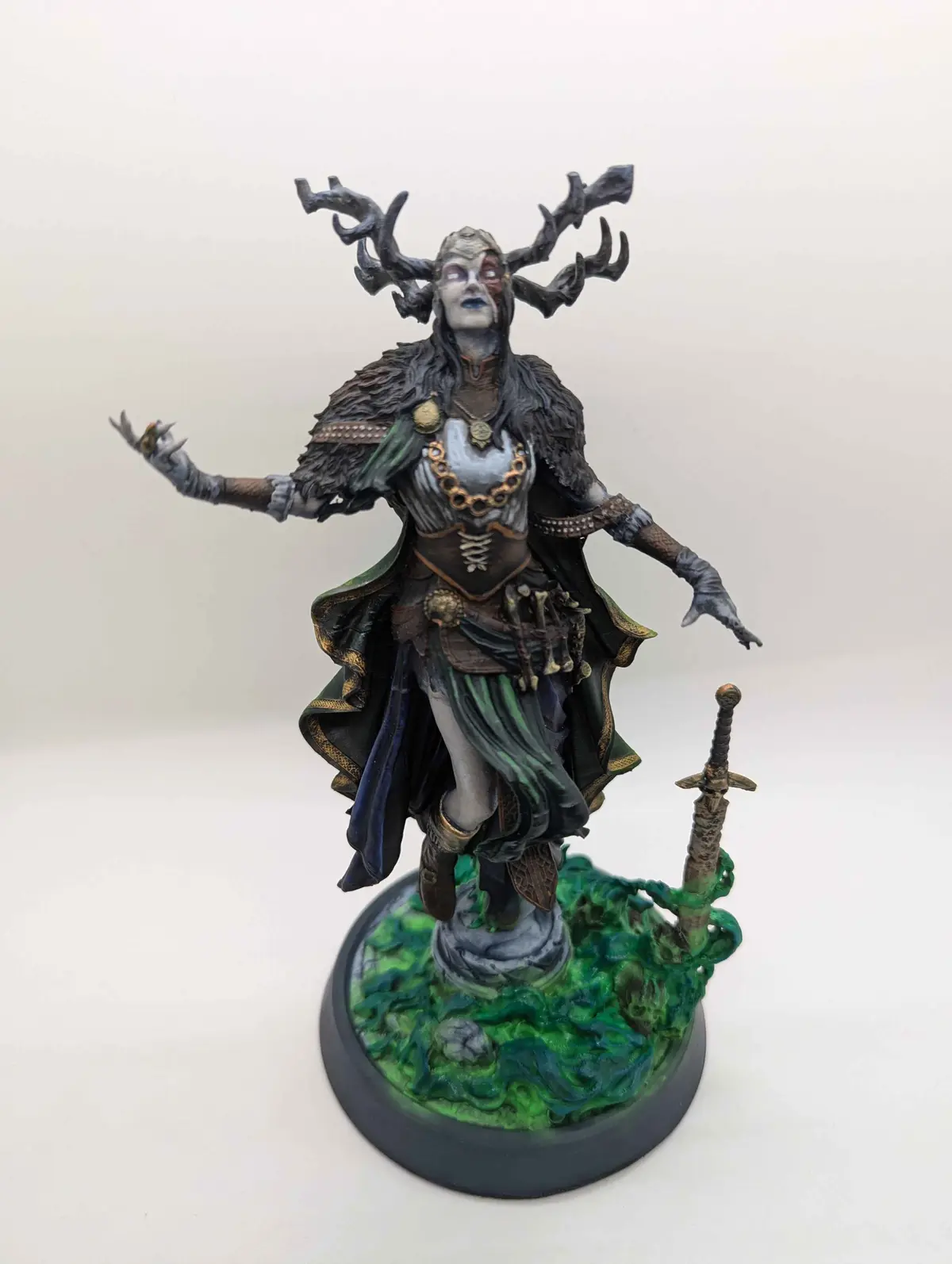 A painted fantasy miniature of a female figure with large, dark antlers, pale skin, and a flowing dark cloak, standing on a…