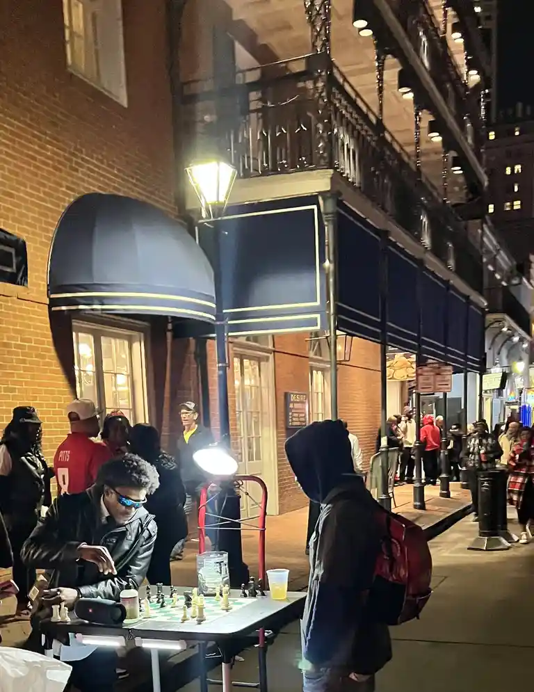 A nighttime street scene shows a man in a black leather jacket and blue-tinted sunglasses intensely focused on a chess game…