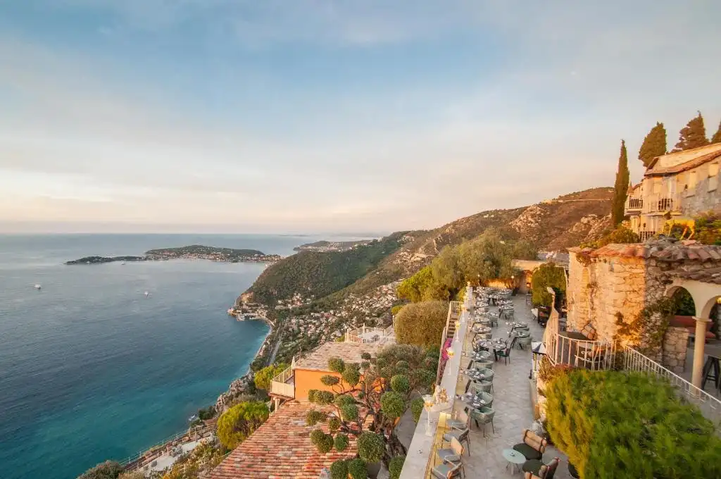 A scenic photo of a cliffside restaurant overlooking the ocean at sunset, with tables and chairs arranged along a stone…
