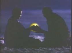 Two silhouetted figures sit facing each other on a beach at night, holding a glowing orb between them. The image is dark and…