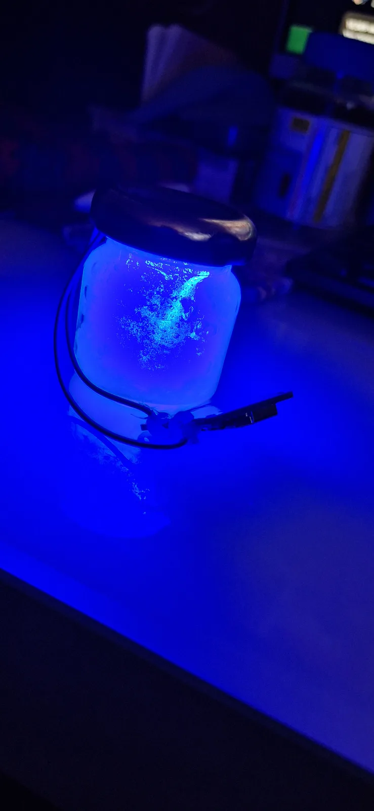 A glass jar with a black lid sits on a surface, glowing with blue light from within, revealing swirling bubbles inside. The…