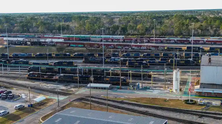 An aerial view of a sprawling rail yard filled with numerous CSX and CN freight trains on multiple tracks, surrounded by…