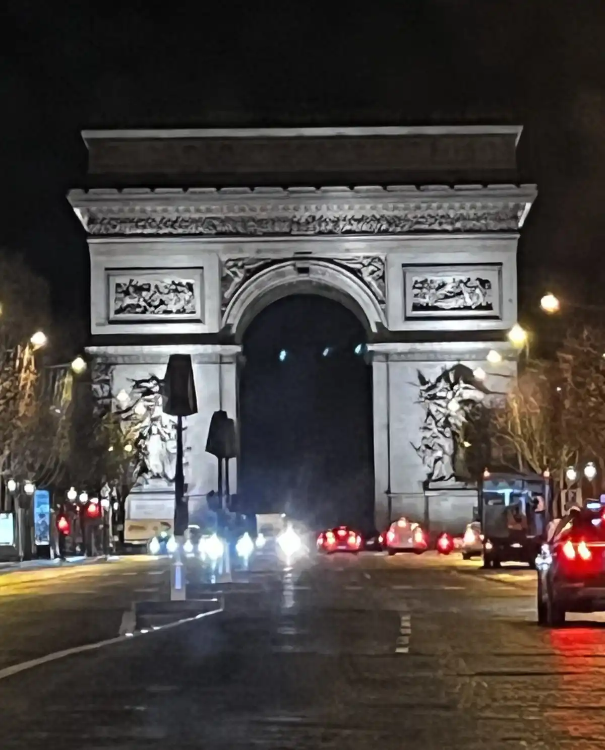 A nighttime photo of the Arc de Triomphe in Paris, with cars' headlights and taillights visible on the wet road. The…