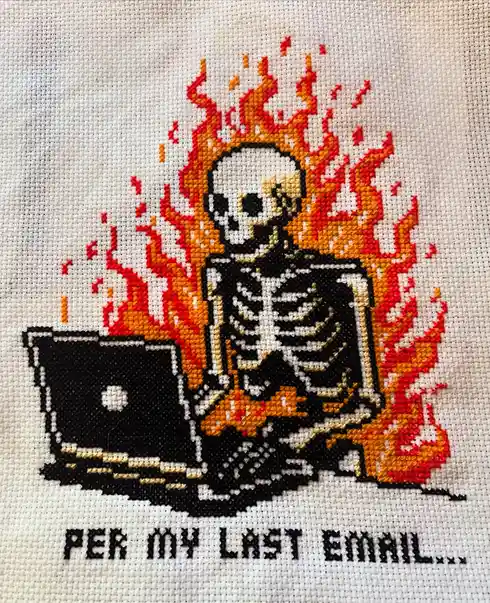 A cross-stitch artwork depicts a flaming skeleton sitting at a laptop, with the text 'PER MY LAST EMAIL...' stitched below…