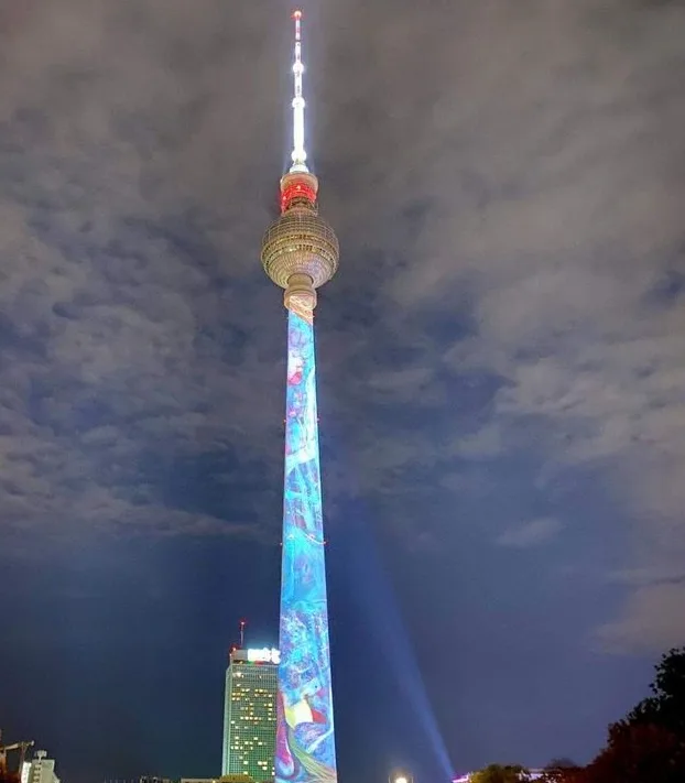 a nighttime photo of the Berlin TV Tower, its main shaft illuminated with a vibrant, colorful projection featuring mermaids…