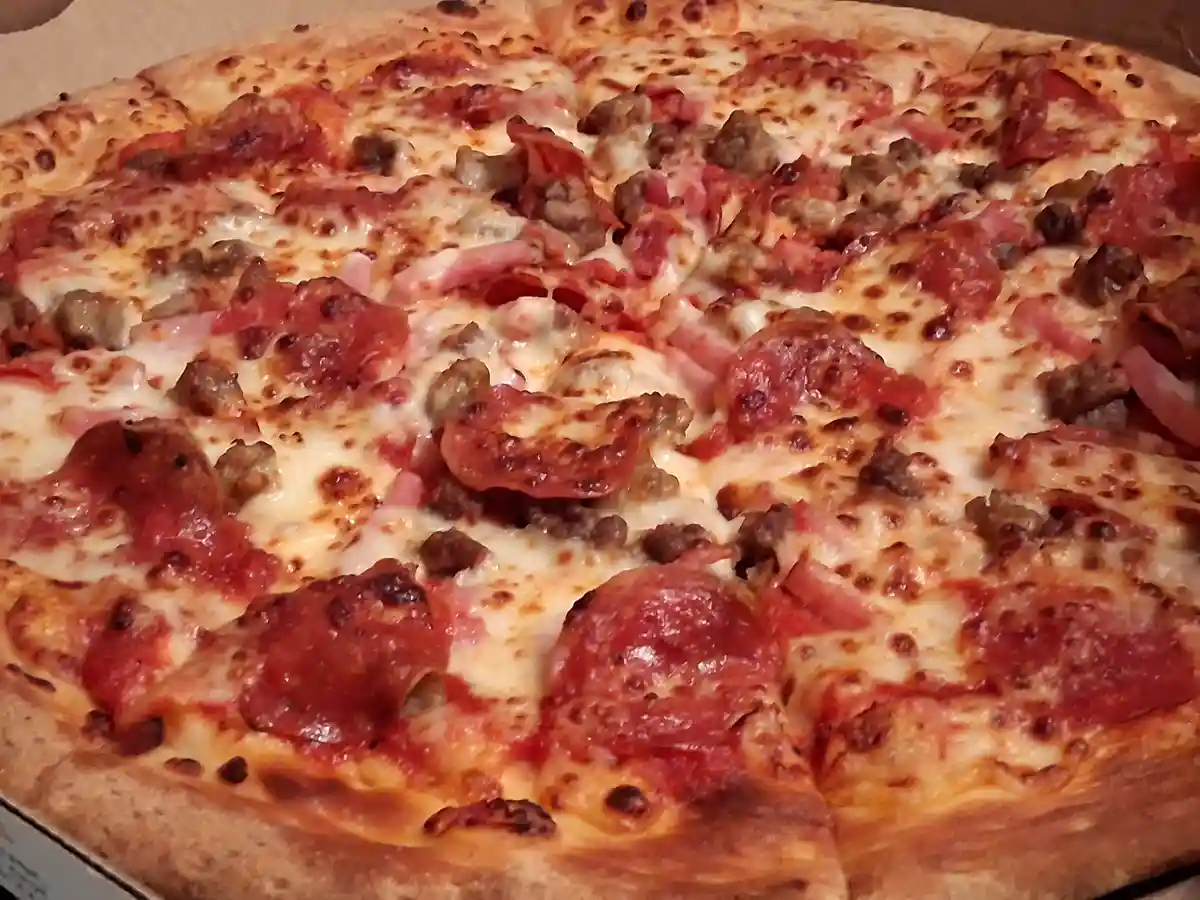 A close-up photo of a freshly baked, sliced pizza in a cardboard box, topped with melted cheese, pepperoni, sausage, and…