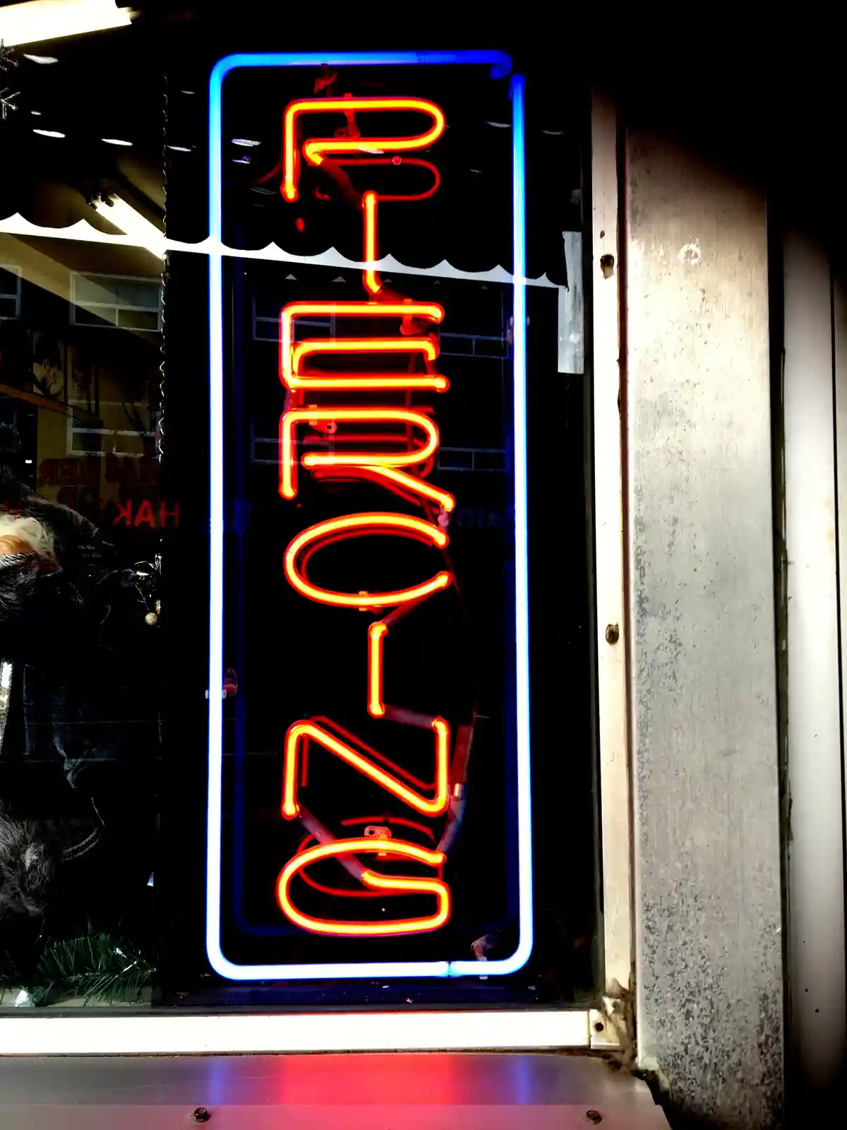 A vertical neon sign with glowing orange-red letters spelling 'OPEN' inside a blue neon border, mounted on a storefront…