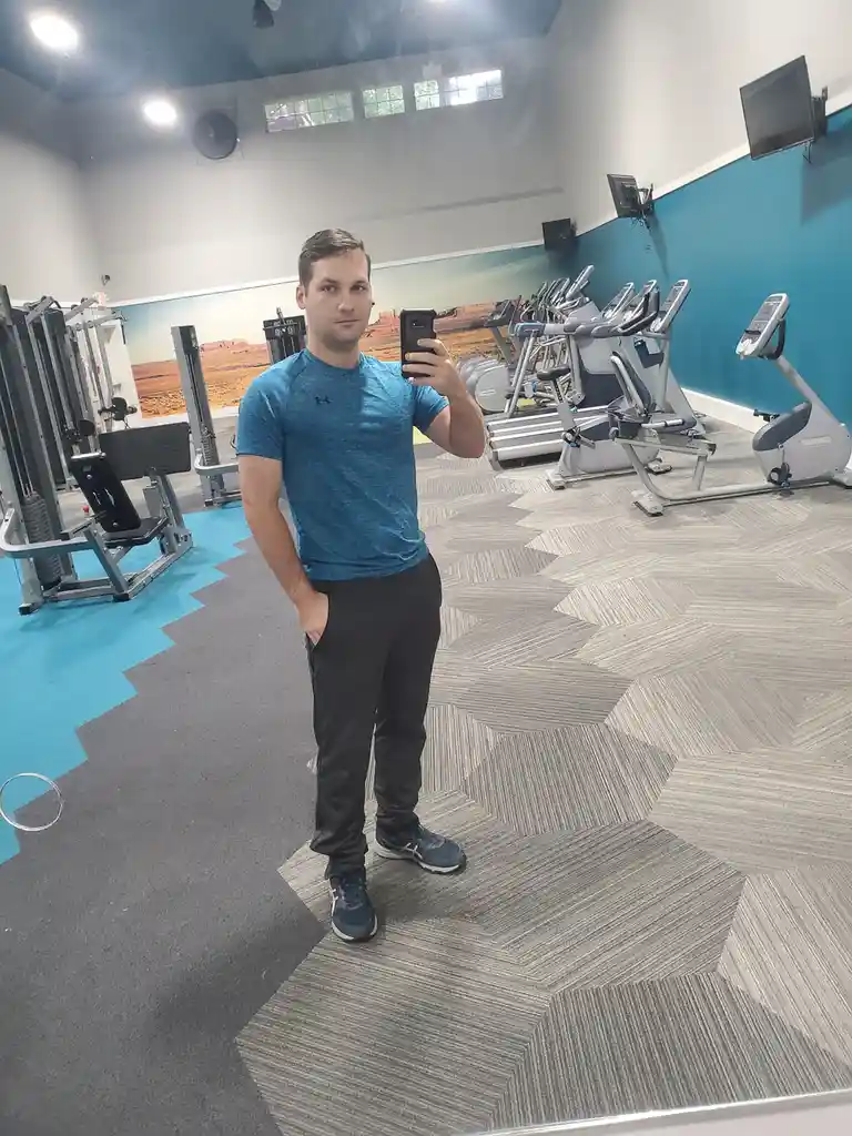A man in a blue Under Armour shirt and black pants takes a mirror selfie in a gym, holding his phone in one hand with the…